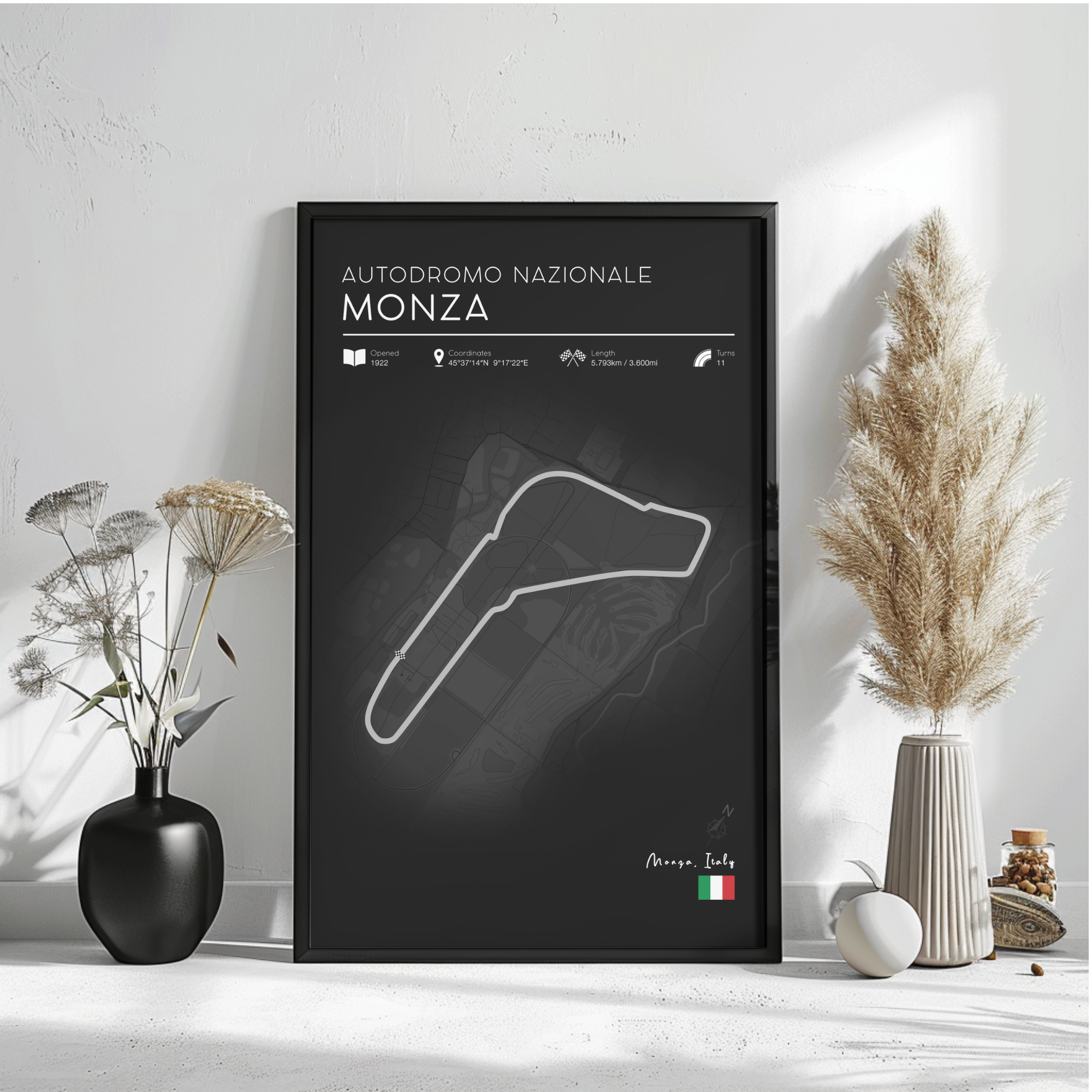 a poster of the Monza racetrack in Italy, with a black background and white text. The poster includes information about the track, such as its name, location, and features. The poster is displayed on a wall, with a vase of flowers and a potted plant to the left, and a small white object to the right.