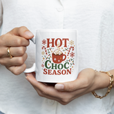 Person holding a mug with 'Hot Choc Season' text and design.