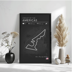 a black poster with a white outline of a race track, titled "CIRCUIT OF THE AMERICAS" and displaying various details about the circuit, including its location, size, and features. The poster is set against a white wall, with a vase of flowers and a potted plant on either side, and a small white object on the floor.