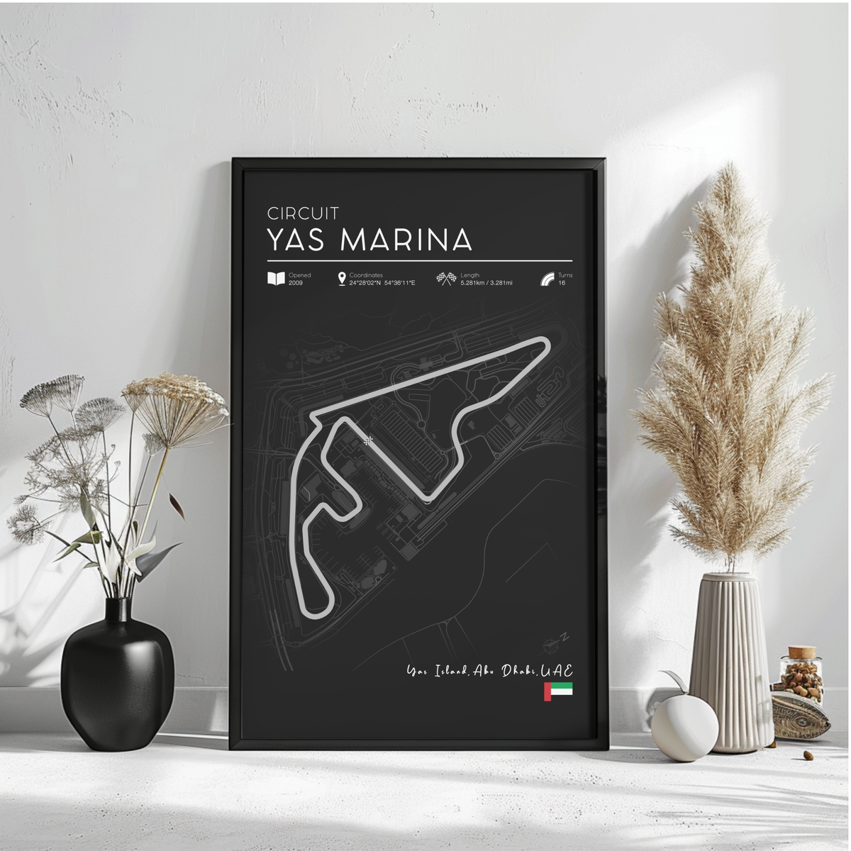 a poster of the Circuit Yas Marina, a racing circuit in Dubai, United Arab Emirates. The poster is displayed on a wall, with a vase of flowers and a potted plant to the left and a small white object to the right.